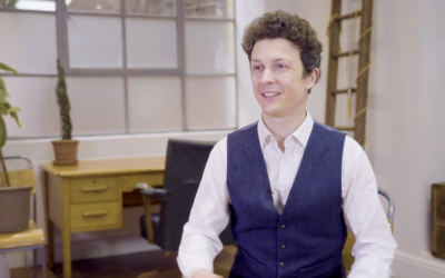 A smiling man with curly hair in a navy waistcoat and white shirt sits in a modern office with wooden furniture, potted plants, and industrial-style windows in the background.