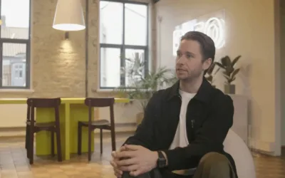 A man in a black jacket and white shirt sits in a modern office space with exposed brick walls, large windows, indoor plants, and a neon sign in the background, speaking in a relaxed setting.