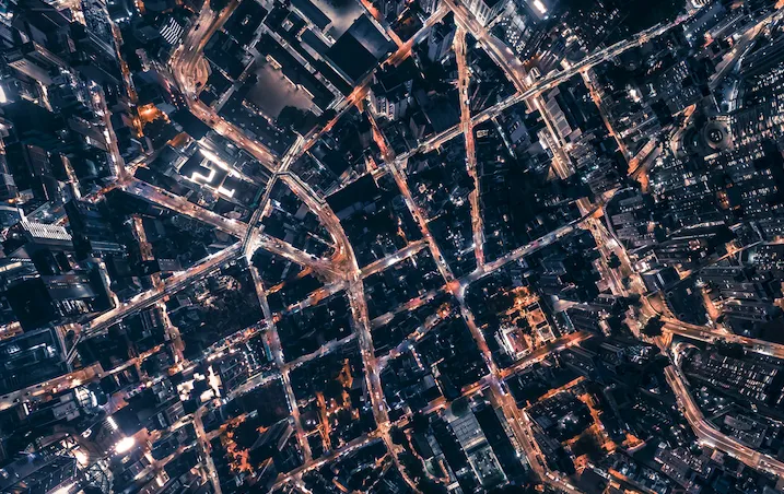 Aerial night view of a dense urban cityscape with illuminated streets, tall skyscrapers, and grid-like road patterns glowing with traffic and building lights.