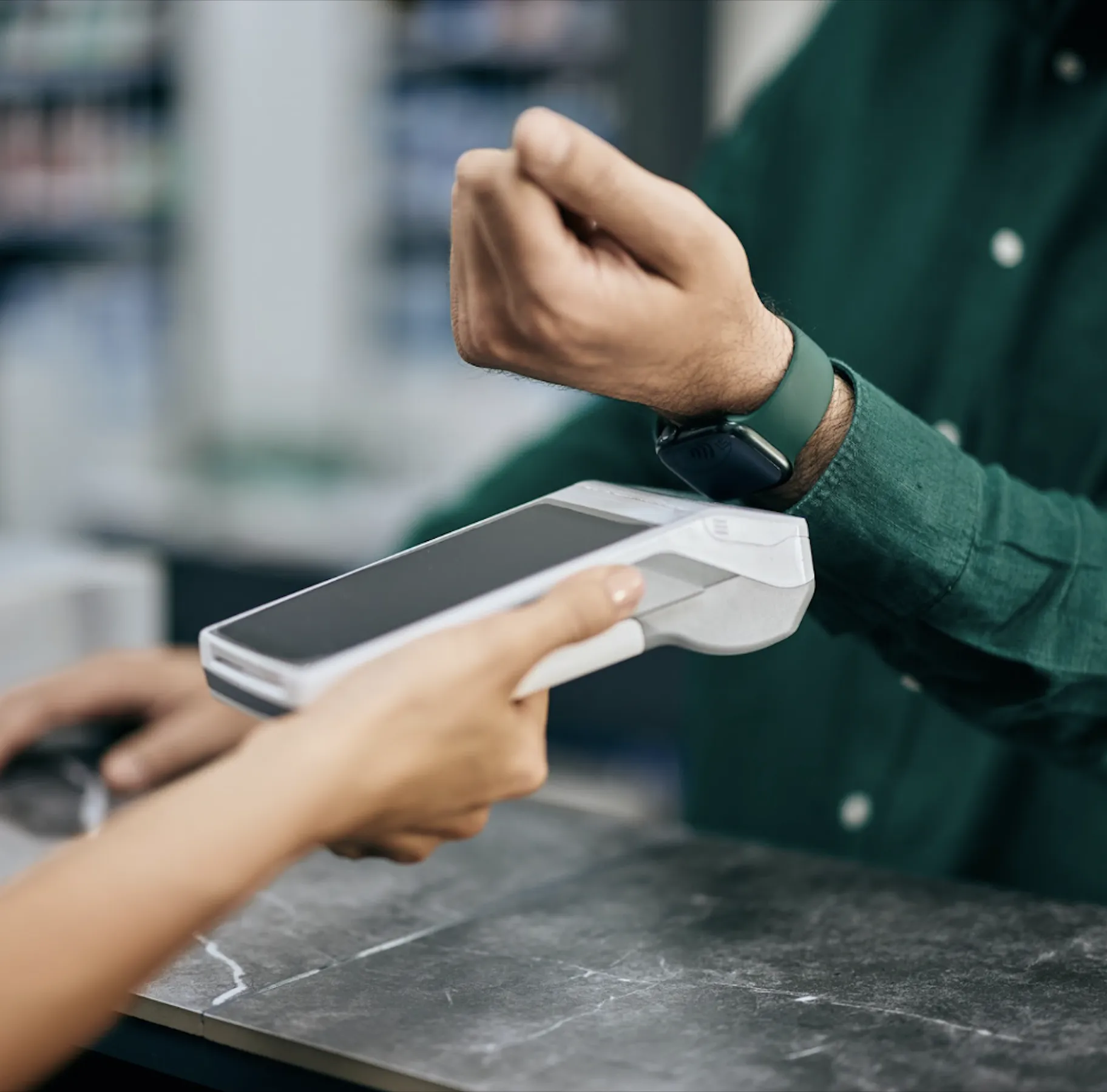 Close-up of a person wearing a smartwatch making a contactless payment at a point-of-sale terminal held by a cashier.