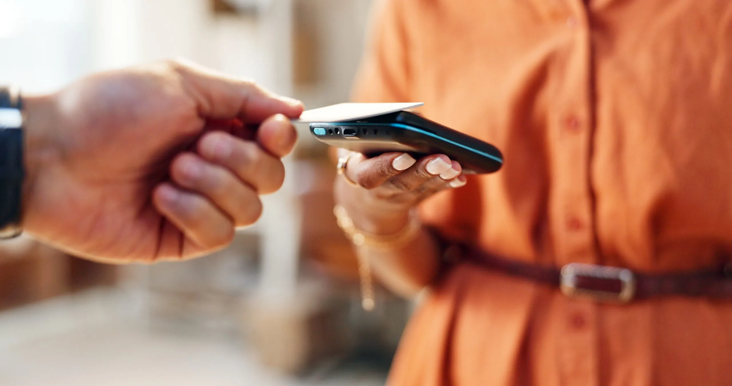 Close-up of a person holding a smartphone while another person makes a contactless payment with a credit card using NFC technology.